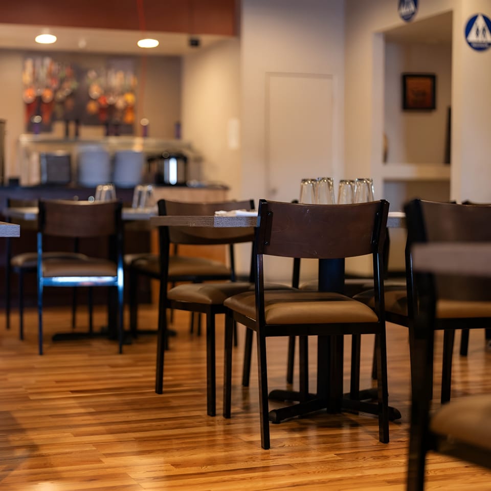 Empty restaurant dining room with dark tables and chairs, and a buffet line in the background.