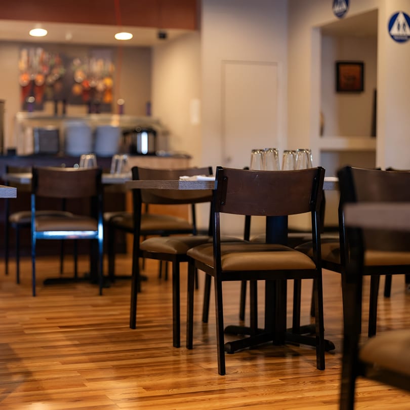 Empty restaurant dining room with dark tables and chairs, and a buffet line in the background.