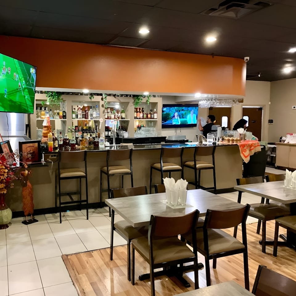 Interior view of a restaurant with a fully stocked bar, dining tables, and two staff members working.
