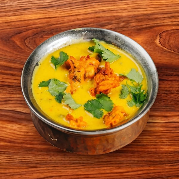 A metal bowl of bright yellow Kadhi Pakora, garnished with fresh green cilantro, on a wooden table.