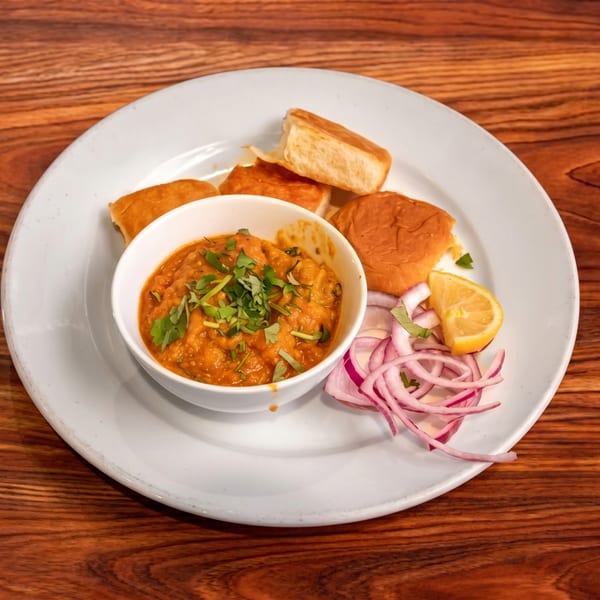 Pav Bhaji, a bowl of vegetable curry with bread rolls, sliced red onions, and a lemon wedge.