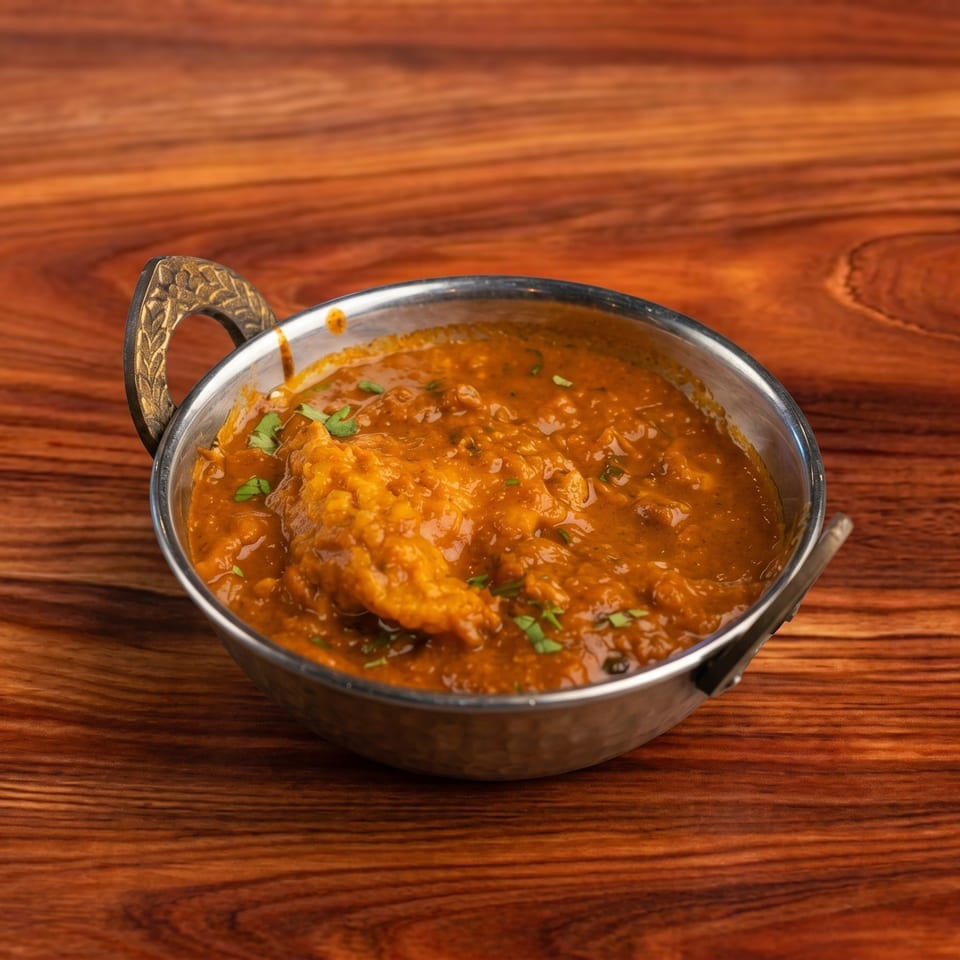 A metal karahi bowl filled with a rich, orange-brown curry, garnished with fresh green herbs.