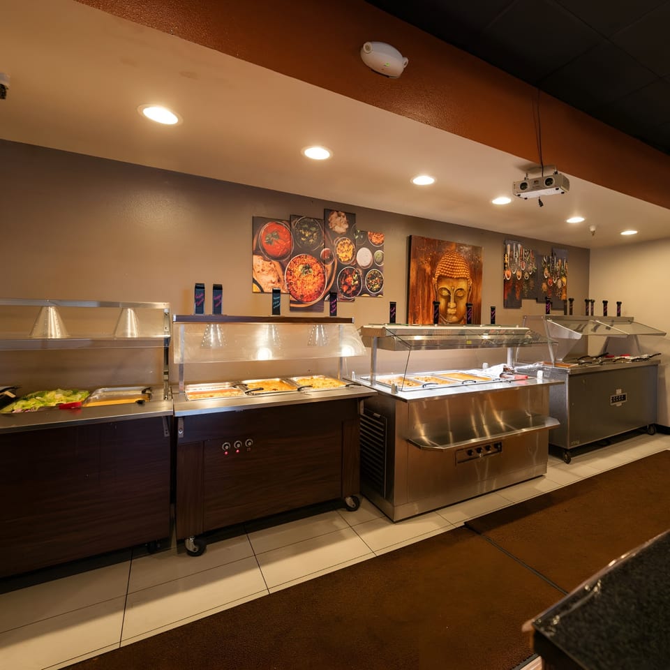 A restaurant buffet line with various food warmers and artistic wall decor, including a Buddha head.