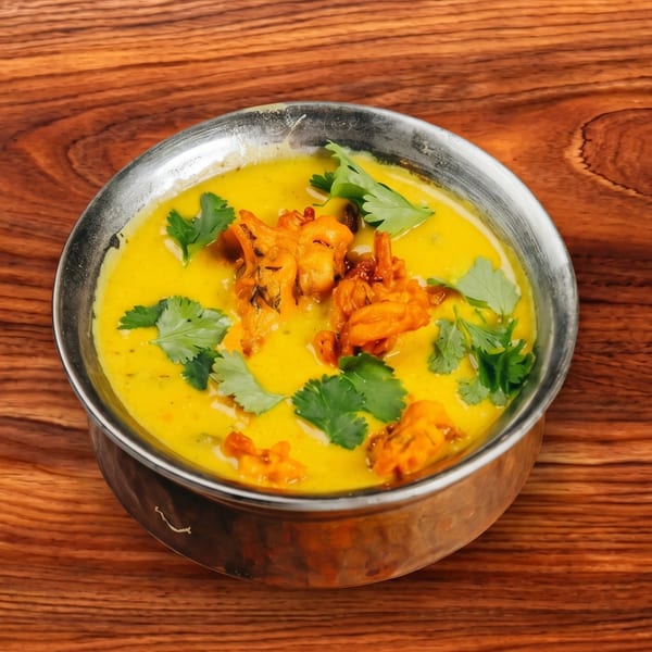 A metal bowl filled with yellow curry, topped with fried fritters and fresh cilantro.