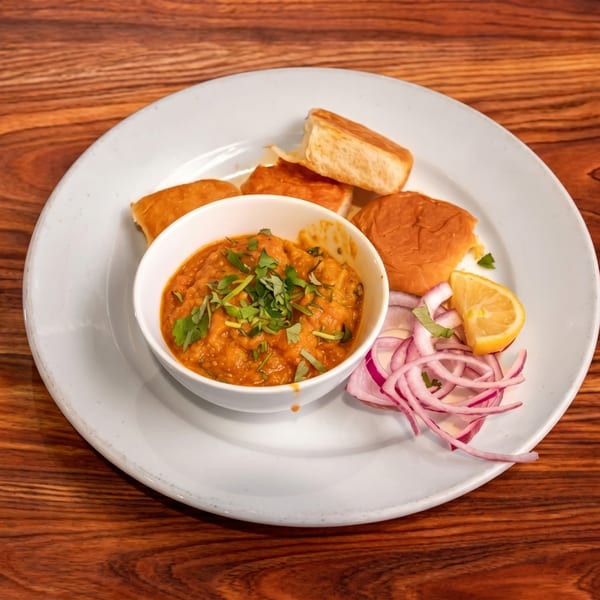 Pav Bhaji, a vibrant orange vegetable curry, with bread, red onions, and lemon on a white plate.